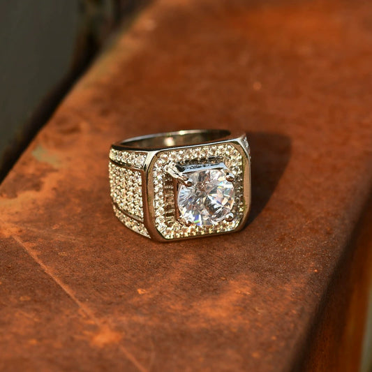 Silver-tone ring with a square face and clear center stone, lying flat on a textured brown surface