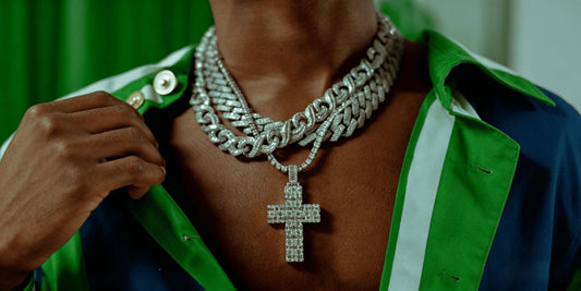 A stylish man displays layers of heavy hip hop jewelry, including a thick Cuban chain and a tennis cross pendant from IceGrind, while adjusting his green and blue shirt collar.