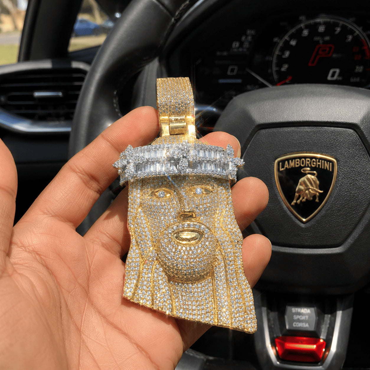 Yellow gold Iced Out Baguette Crown Jesus Piece held in hand against a luxury car steering wheel.
