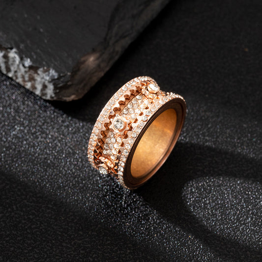Rose-gold-tone wide ring with stone rows and a gear-like center, lying flat on a dark textured surface