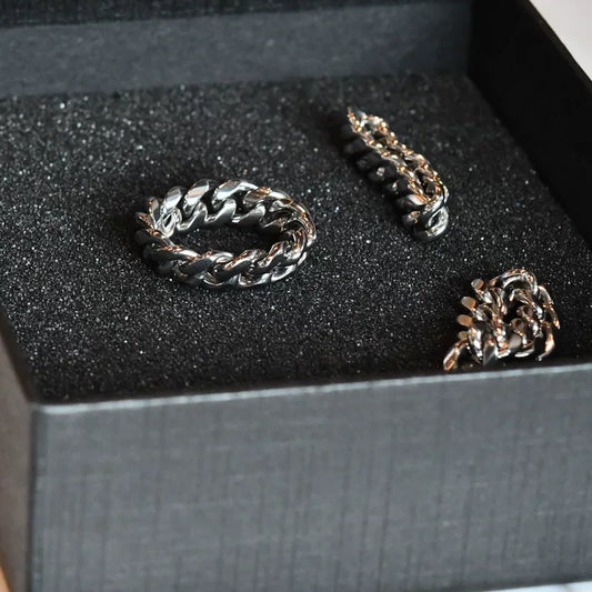 A silver-tone Cuban link ring lies flat in a black box, with two other metallic items nearby