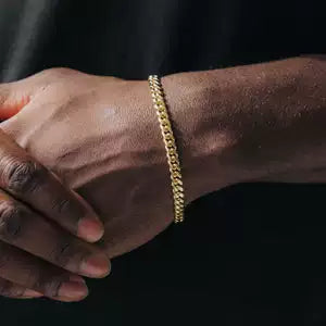 Gold-tone Cuban link bracelet worn on a wrist with both hands visible against black