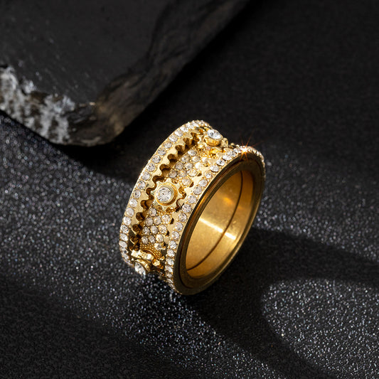 Gold-tone sculpted ring with gear-like designs and pavé-set stones, lying flat beside a dark rock