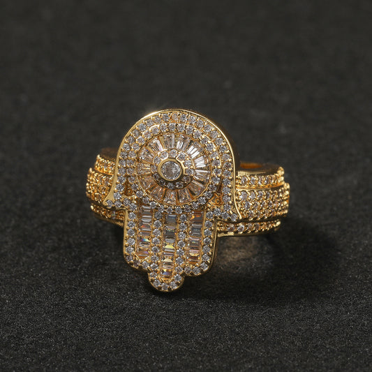 Gold-tone sculpted ring with a circular stone center and hand-shaped motif, lying flat on a dark surface